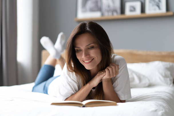Mujer joven acostada en la cama y leyendo un libro en casa. - Foto, imagen