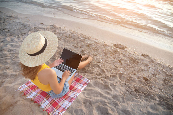 Young woman freelancer in denim shorts and a straw hat with a laptop on her legs sits on the sandy seashore at sunset. Remote work via the Internet in a warm effort by the ocean - Foto, Imagen