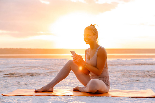 jonge blanke vrouw houden mobiele telefoon terwijl het doen van workout op fitness mat buiten - Foto, afbeelding