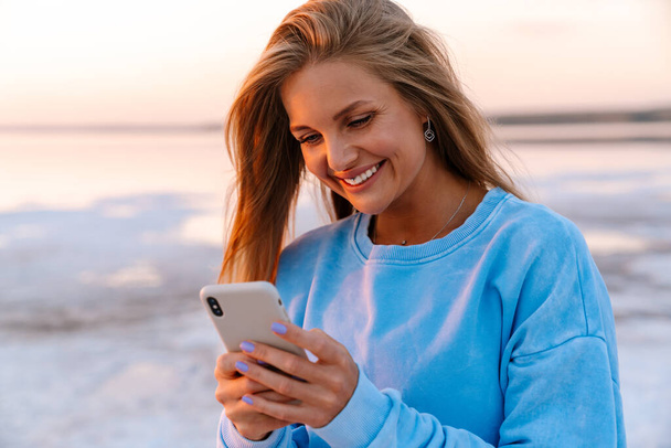 młody biały szczęśliwy blondynka kobieta uśmiech i trzymając telefon komórkowy na zewnątrz - Zdjęcie, obraz