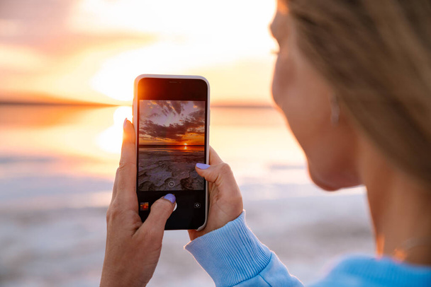 Young caucasian happy blonde woman taking photo of sunset on cellphone outdoors - Фото, зображення