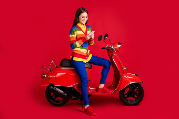 Photo portrait of woman texting holding phone in two hands sitting on scooter isolated on vivid red colored background - Фото, изображение