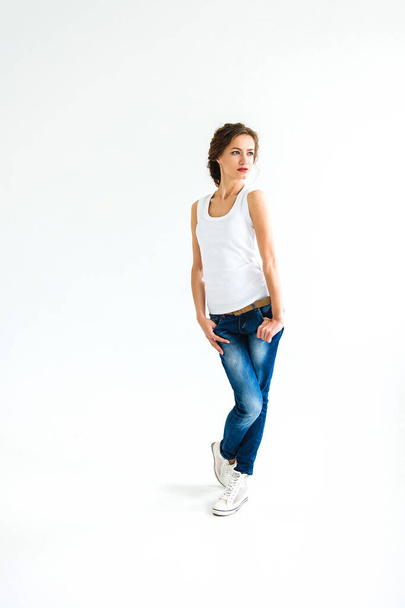 alegre chica en una camiseta blanca y pantalones vaqueros azul oscuro en el estudio sobre un fondo blanco se levanta, se sienta, corre - Foto, Imagen