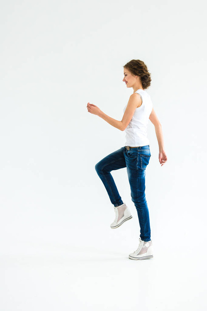 fröhliches Mädchen in weißem T-Shirt und dunkelblauer Jeans im Studio vor weißem Hintergrund steht, sitzt, rennt - Foto, Bild