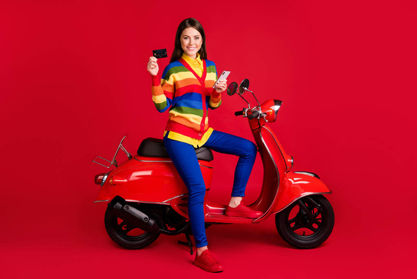 Photo portrait of woman holding phone credit card in hands sitting on retro scooter isolated on vivid red colored background - Photo, Image