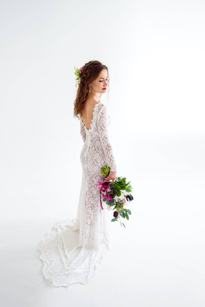 joyeuse jeune mariée dans une robe blanche tricotée posant avec un bouquet de fleurs dans le studio sur un fond blanc - Photo, image