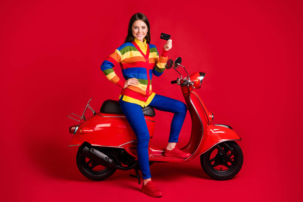 Photo portrait of confident woman holding plastic card in one hand sitting on scooter isolated on vivid red colored background - Foto, Imagem