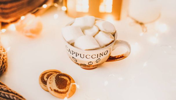 Coffee cup with marshmallows and cookies on bokeh lights background. Christmas and New Year cozy still life lights and decorations - Fotó, kép
