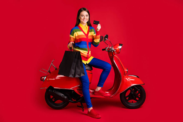 Photo portrait of smiling woman holding credit card shopping bags sitting on scooter isolated on vivid red colored background - Photo, Image