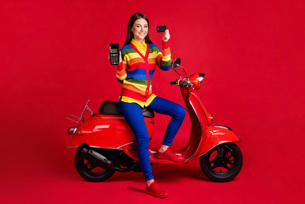 Photo portrait of happy woman holding credit card terminal in hands sitting on scooter isolated on vivid red colored background - Photo, image