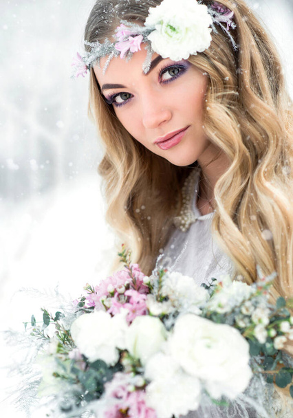 Attractive winter woman in a white snow forest with a beautiful flower wreath and bouquet. Bride makeup and hairstyle - Fotó, kép