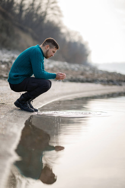 A young man in a turquoise sweater wets his hands in the river - Photo, image