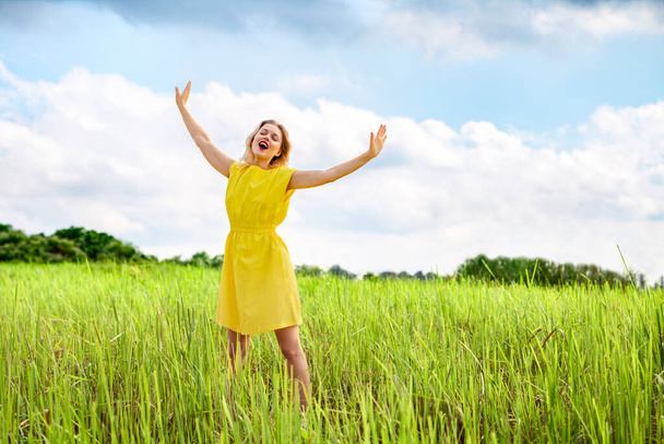 Happy young woman in yellow dress is singing with closed eyes and raising her hands on green meadow. Enjoy summertime. Open your sound voise. - Foto, imagen