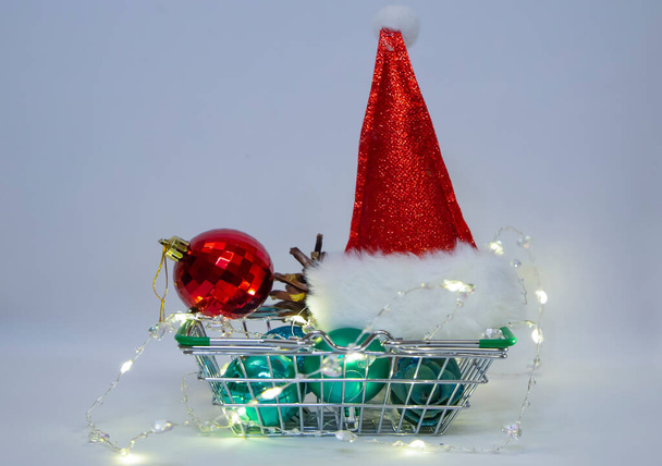 Merry Christmas and Happy New year:Santa Claus hat Christmas balls and electric garland in shopping basket - Foto, Imagem