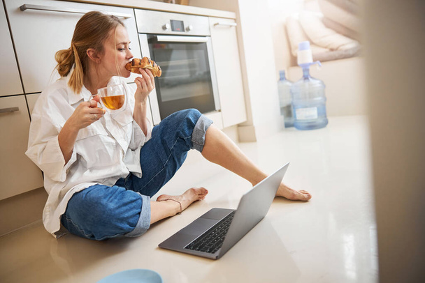 Mulher bonita comendo croissant e usando laptop em casa - Foto, Imagem