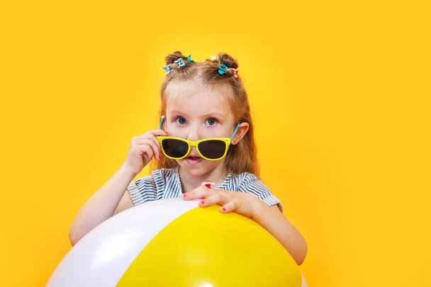 Funny happy child in sunglasses with summer beachball on yellow background - Foto, Imagem