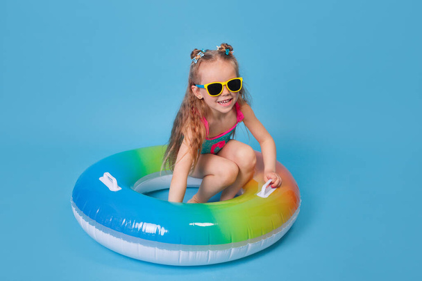 Happy child girl in swimsuit and sunglasses with swimming ring on blue background - 写真・画像