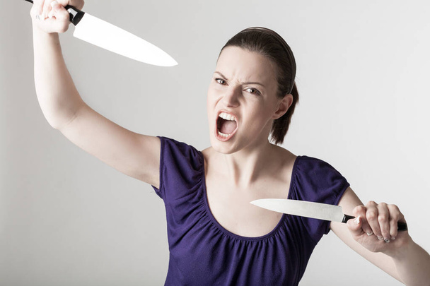Μια νεαρή γυναίκα που ουρλιάζει κάνει χειρονομίες με δύο κοφτερά μαχαίρια κουζίνας.  - Φωτογραφία, εικόνα