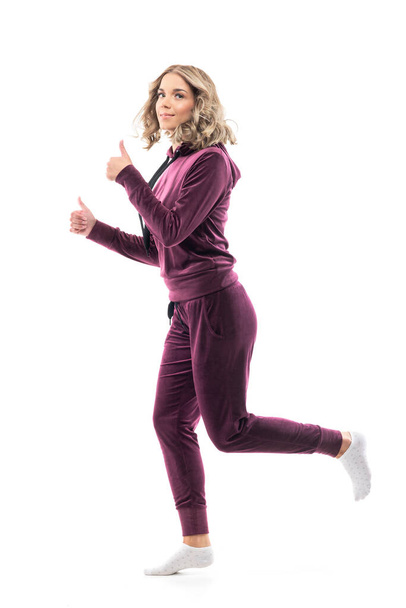 Vista lateral da mulher de beleza feliz em casa usar sweatsuit correndo e mostrando os polegares para cima. Comprimento total do corpo isolado no fundo branco.  - Foto, Imagem