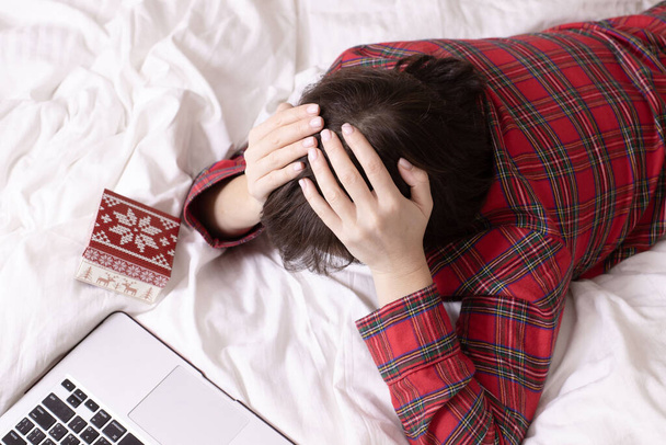 Concept of psychological problems, New year on self-isolation, depression, loneliness during the holidays. Woman in christmas pajamas grabbed her head lying face down on white bed near gift, laptop. - Photo, Image