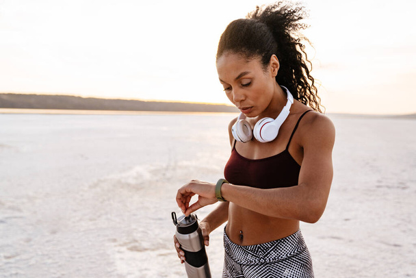 Imagen de una joven deportista afroamericana mirando smartwatch mientras hace ejercicio sobre la naturaleza - Foto, imagen
