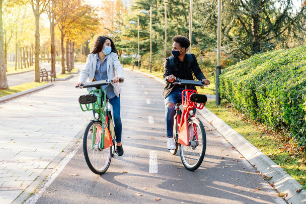 front of two young man and woman riding along a bicycle path with a sharing electric bicycle in a beautiful park with many trees at sunset wearing a face mask for the 2020 covid19 coronavirus pandemic - Zdjęcie, obraz