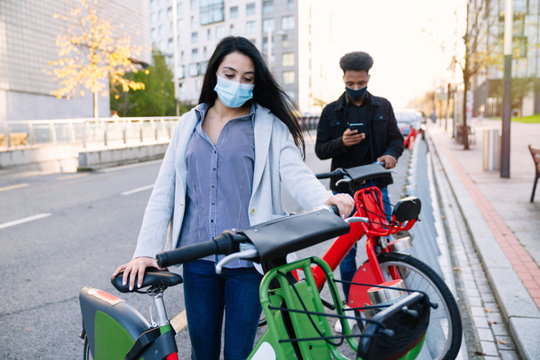 Молодой марокканец с сотовым телефоном и кавказская девушка берут напрокат электрический велосипед с парковки для велосипедов и надевают маску для лица во время пандемии коронавируса 2020 года - Фото, изображение