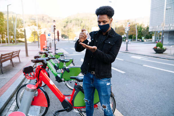 路上自転車の駐車場でレンタルした電気自転車を拾うために若いモロッコ人が手に水道アルコールジェルを注いでおり、 covid19コロナウイルスパンデミックの顔マスクを着ています - 写真・画像