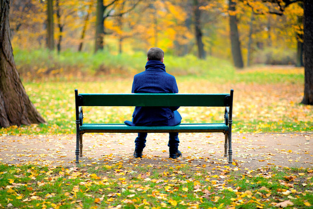Μοναχικός άντρας κάθεται σε ξύλινο παγκάκι σε πάρκο το φθινόπωρο. - Φωτογραφία, εικόνα