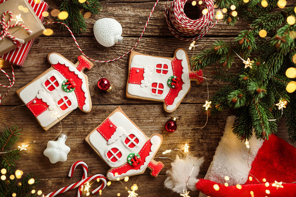 Kerst peperkoek huizen, sparren takken, rode decoraties, Santa hoed, snoep, lichten slinger op houten achtergrond. Vrolijk kerstfeest en nieuwjaarsvakantie. Kerstkoekjes, bovenaanzicht, bokeh - Foto, afbeelding