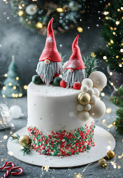 Christmas cake decorated with sweet figures Santa's gnomes, fir branches and bells on bright background with garland, candy, bokeh. Winter baking at Xmas or New Year holiday, close up - Fotografie, Obrázek