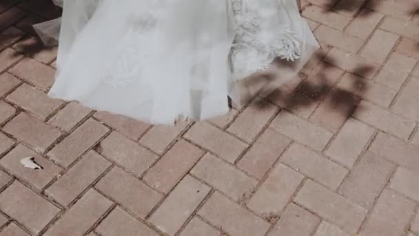 La mariée avec un long voile blanc de fatin et de dentelle chère va de l'avant et tire le voile sur les tuiles dans la cour par une journée ensoleillée et sur le fond d'ombres des feuilles des arbres - Séquence, vidéo
