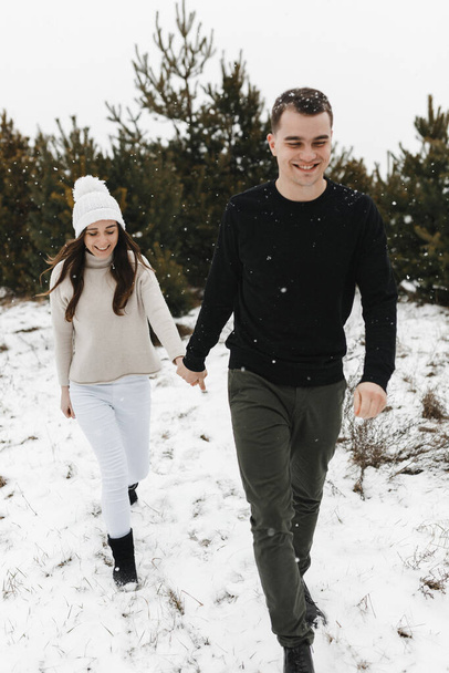 Young man and woman going on the snow in the pine forest holding each other hands. Love, relationship, winter holidays. Winter couple photo ideas - Photo, Image