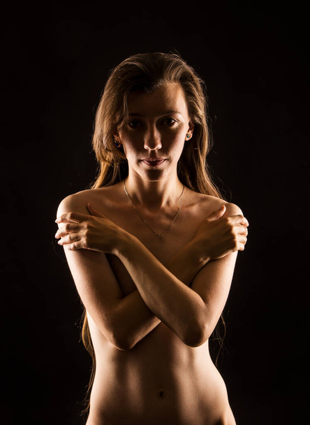 Portret van een naakte jonge vrouw met lang haar in contrast met licht op een donkere achtergrond. Studioshoot. - Foto, afbeelding