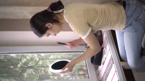 Een jonge vrouw voor een raam kijkt in een kleine spiegel en brengt make-up aan met een borstel. Zorgen voor de huid. Verticale video. - Video