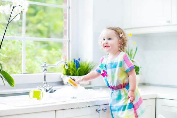 χαριτωμένο νήπιο σγουρά κορίτσι σε ένα πολύχρωμο φόρεμα πλύσιμο των πιάτων - Φωτογραφία, εικόνα
