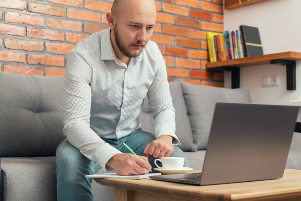 Бородатый лысый мужчина, бизнесмен или фрилансер, сидящий на диване с ручкой и блокнотом в руке, работающий на ноутбуке из дома, современный дизайн интерьера лофта. - Фото, изображение