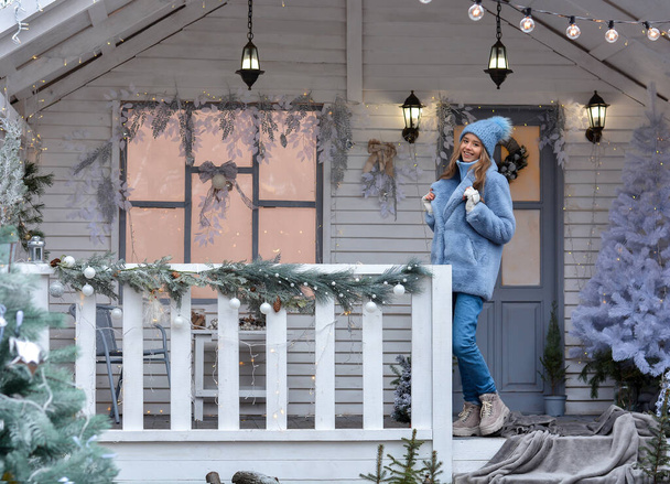 Ένα χαρούμενο κορίτσι μοντέλο με βαθύ καπέλο, γούνινο παλτό, γάντια στέκεται στη βεράντα του χριστουγεννιάτικου σπιτιού! Ευτυχισμένο το Νέο Έτος και Καλά Χριστούγεννα! - Φωτογραφία, εικόνα
