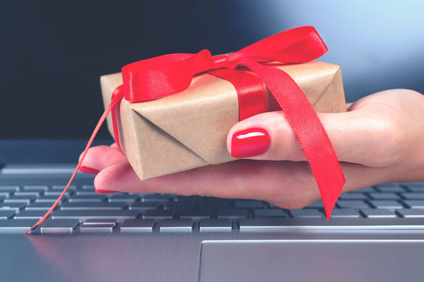 Woman's hand holding present box with red bow and laptoptop computer as background behind it for your project. Seasonal Christmas sale, black friday, online shopping and cyber monday concept - Photo, Image