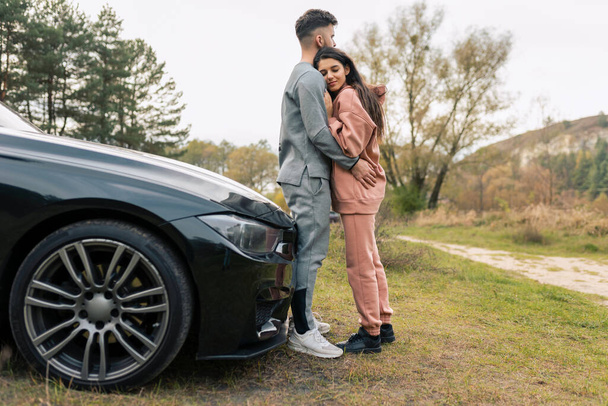 Eşofman giymiş romantik bir çift, arabanın yanında dururken sarılıyor.  - Fotoğraf, Görsel