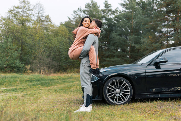Bella giovane coppia in tuta da ginnastica vicino alla loro auto. Felicità ragazza tra le braccia del suo fidanzato - Foto, immagini