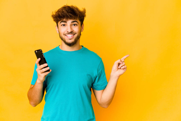 Young indian man holding a phone smiling and pointing aside, showing something at blank space. - Φωτογραφία, εικόνα