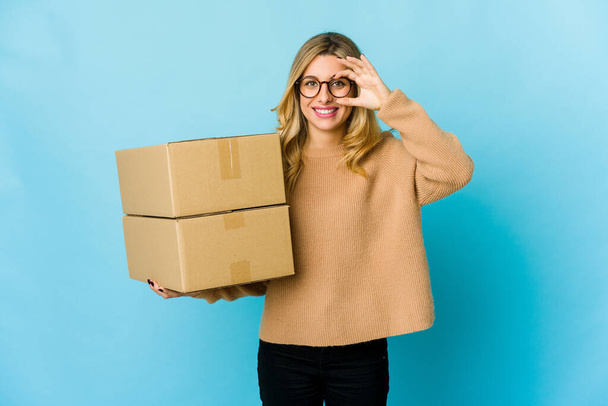Νεαρή ξανθιά καυκάσια γυναίκα κρατώντας κουτιά για να κινηθεί ενθουσιασμένος κρατώντας ok χειρονομία στο μάτι. - Φωτογραφία, εικόνα