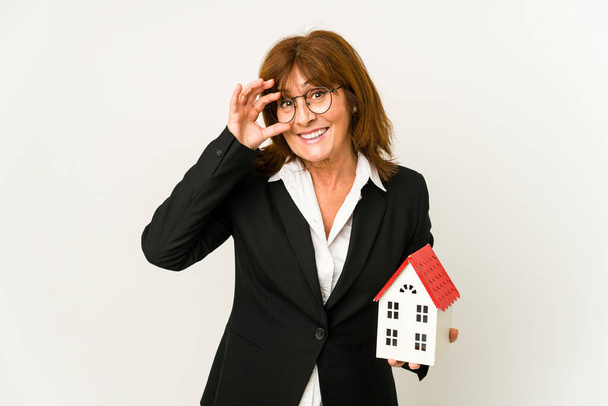 Agent nieruchomości w średnim wieku trzyma model domu odizolowany podekscytowany trzymając ok gest na oku. - Zdjęcie, obraz