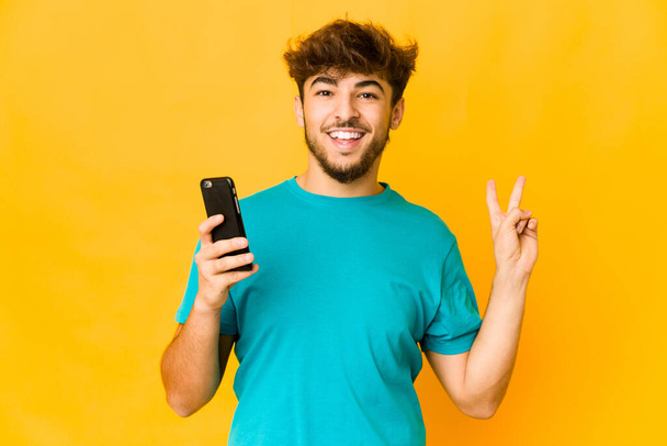 Young indian man holding a phone joyful and carefree showing a peace symbol with fingers. - Φωτογραφία, εικόνα