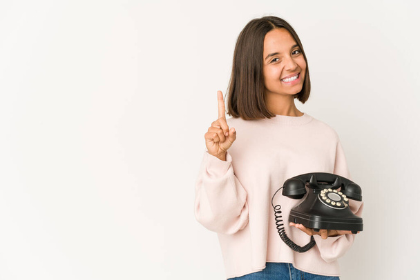 Junge hispanische Frau hält ein altes Telefon in der Hand und zeigt die Nummer eins mit dem Finger. - Foto, Bild