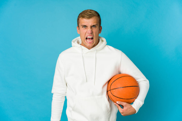 Junge kaukasische Mann spielt Basketball isoliert auf blauem Hintergrund schreit sehr wütend und aggressiv. - Foto, Bild