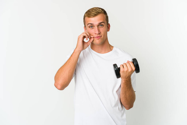 Young caucasian man holding a weight isolated on white background with fingers on lips keeping a secret. - 写真・画像