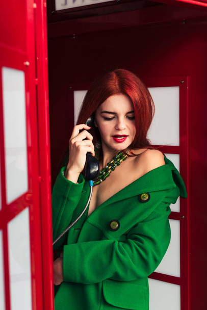 menina ruiva em um casaco verde falando no telefone em uma cabine de telefone vermelho - Foto, Imagem