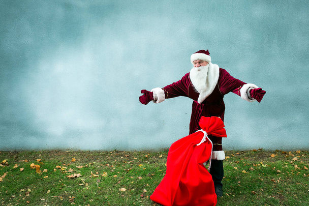 Der Weihnachtsmann mit einem großen Sack voller Geschenke. Der Weihnachtsmann wirft die Hände in die Höhe und zeigt, was für eine große Tüte. Nahaufnahme auf grauem Hintergrund und grünem Gras. - Foto, Bild
